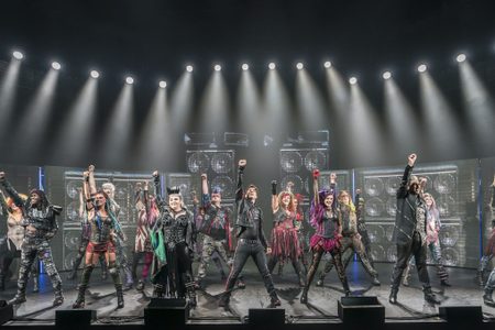 A group of We Will Rock You performers stand on stage with one hand in the air in a rockstar-like stance