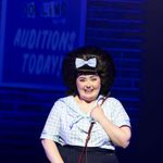 Hairspray Production photo. Tracey Turnblad is stood in front of a poster for The Corny Collins Show. She is holding a red school bag and looks excited.