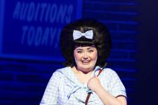 Hairspray Production photo. Tracey Turnblad is stood in front of a poster for The Corny Collins Show. She is holding a red school bag and looks excited.