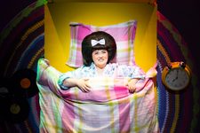 Hairspray Production photo. Tracey Turnblad is lying in a brightly coloured bed. There are some vinyl records and a clock by her side.