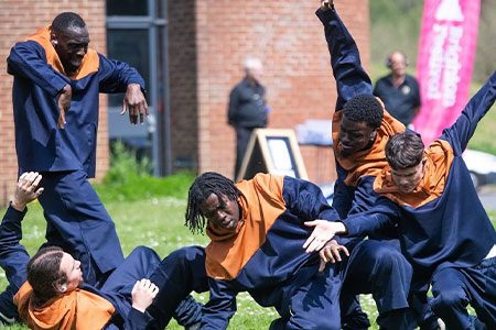 Closer to my dreams Weekender image. Four young dancers wearing orange and blue jumpers are leaning towards the floor, they look like they are all reaching for something far away.