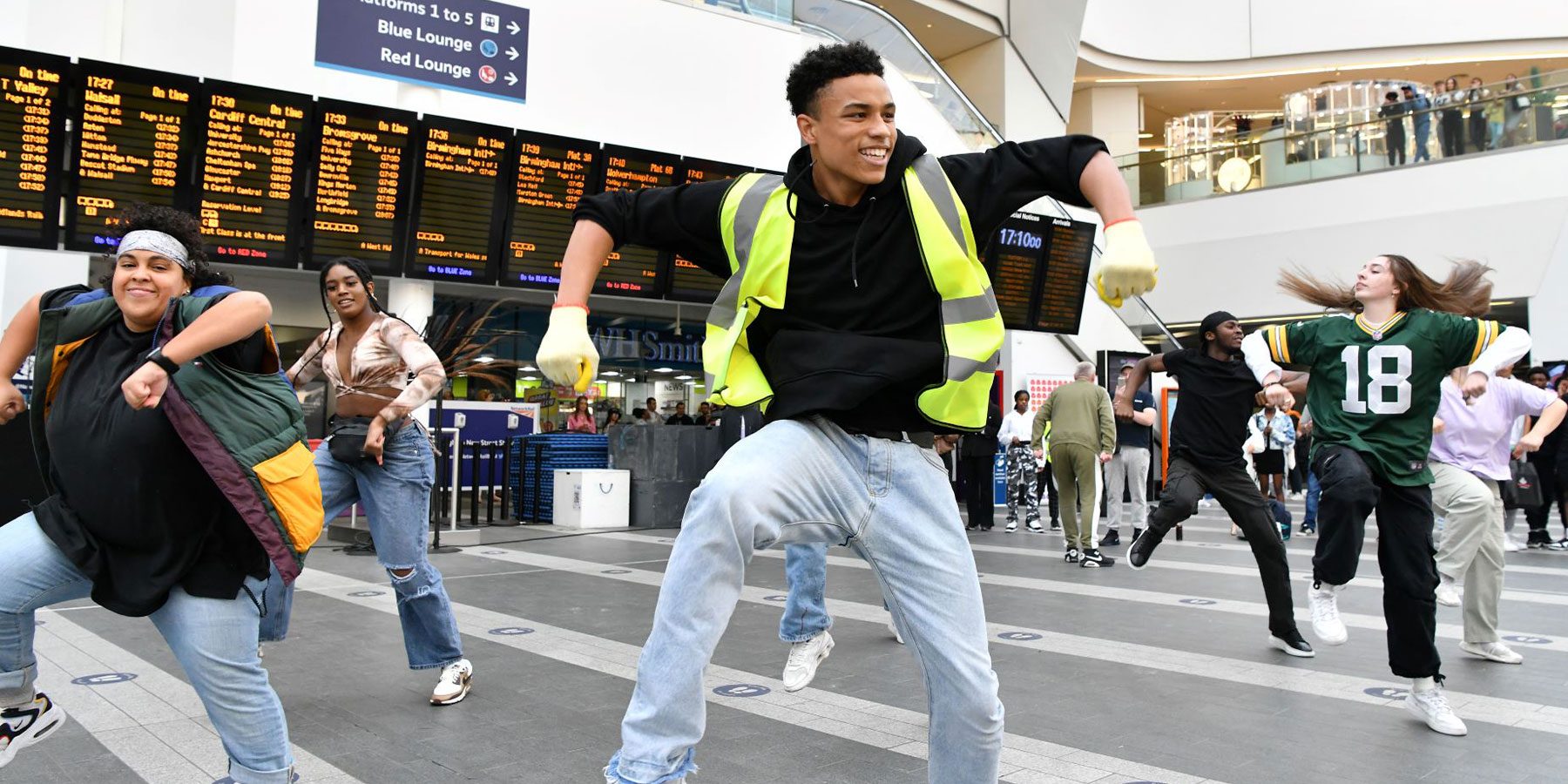 Dance Flashmob Weekender image. Approximately 7 dancers are in Newhall Street station, dancing a fun hip-hop style dance in front of the train times.