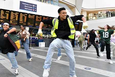 Dance Flashmob Weekender image. Approximately 7 dancers are in Newhall Street station, dancing a fun hip-hop style dance in front of the train times.