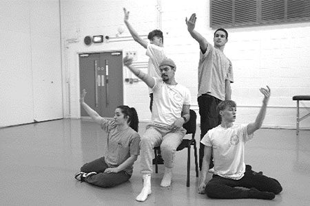 Swan Break Weekender image. A black and white picture of 5 dancers, two sat on the floor and 2 standing around 1 on a chair. They all have their arms outstretched in symmetry with each other.