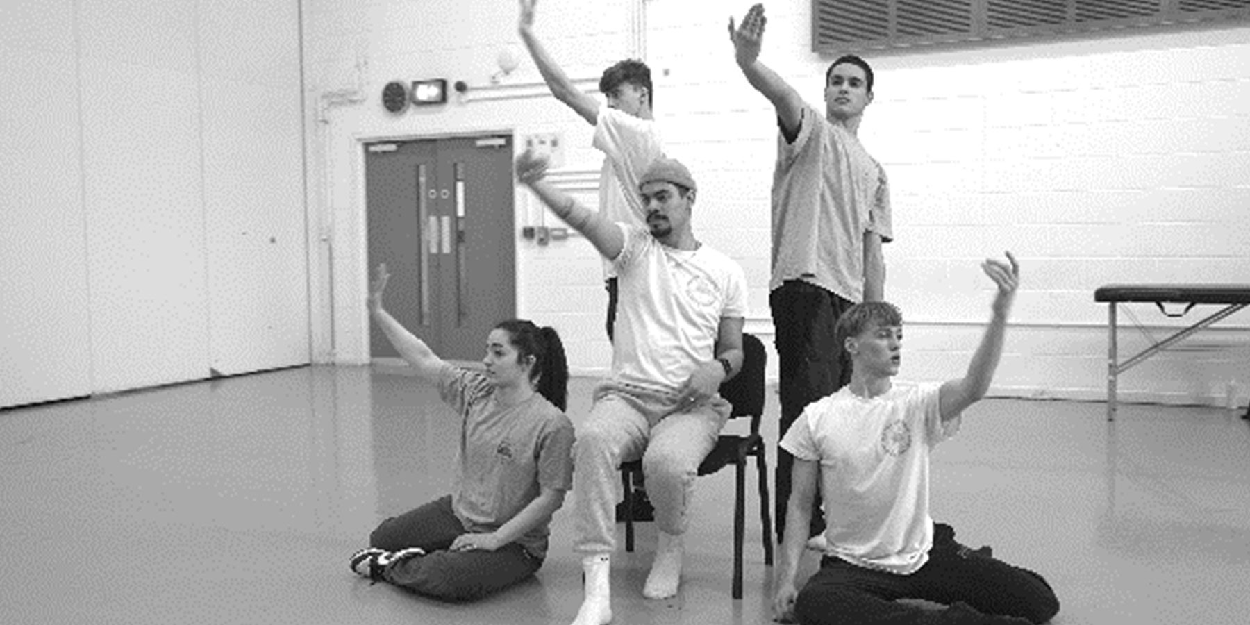 Swan Break Weekender image. A black and white picture of 5 dancers, two sat on the floor and 2 standing around 1 on a chair. They all have their arms outstretched in symmetry with each other.