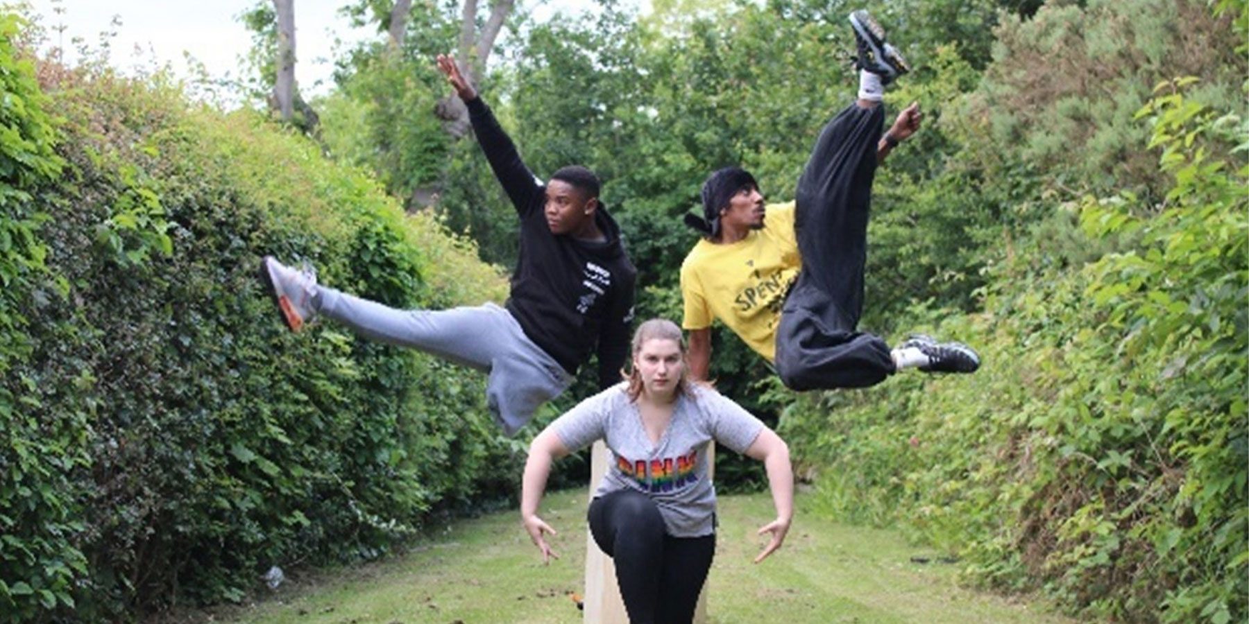 Synergy Weekender image. Three dancers are stood in lots of greenery. One is bent forwards with her arms rounded, and the other two are jumping either side of her with their legs in the air.