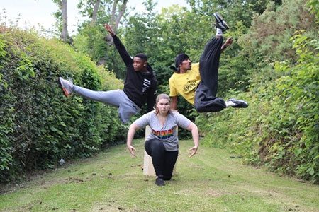 Synergy Weekender image. Three dancers are stood in lots of greenery. One is bent forwards with her arms rounded, and the other two are jumping either side of her with their legs in the air.