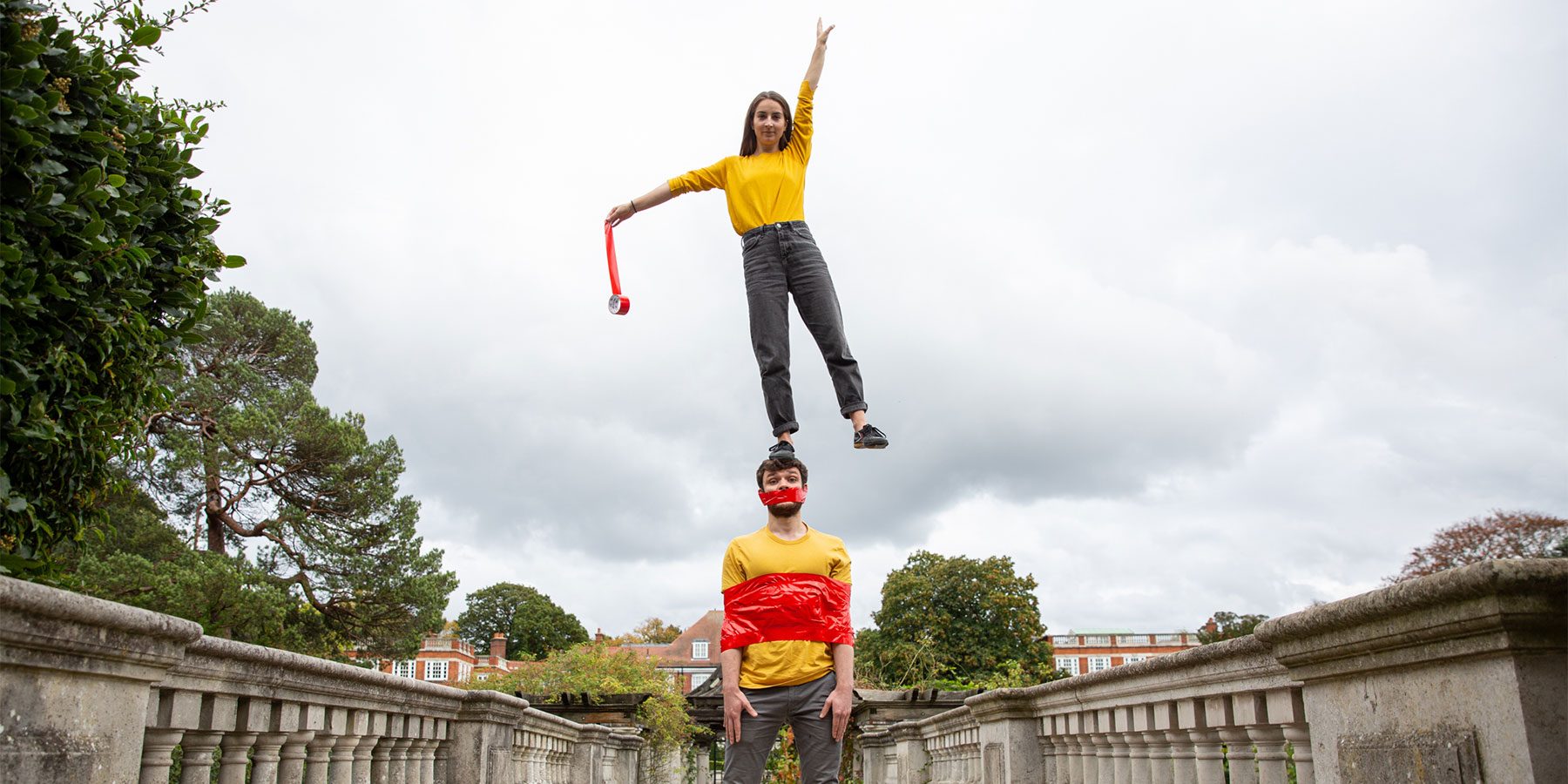 Tape That Weekender image. Two acrobats re on a bridge, one is stood on the others' head. The dancer on the head is holding a roll of red tape, and the dancer on the ground is tied up by the red tape.