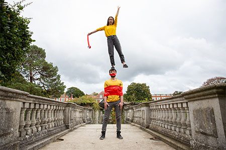 Tape That Weekender image. Two acrobats re on a bridge, one is stood on the others' head. The dancer on the head is holding a roll of red tape, and the dancer on the ground is tied up by the red tape.