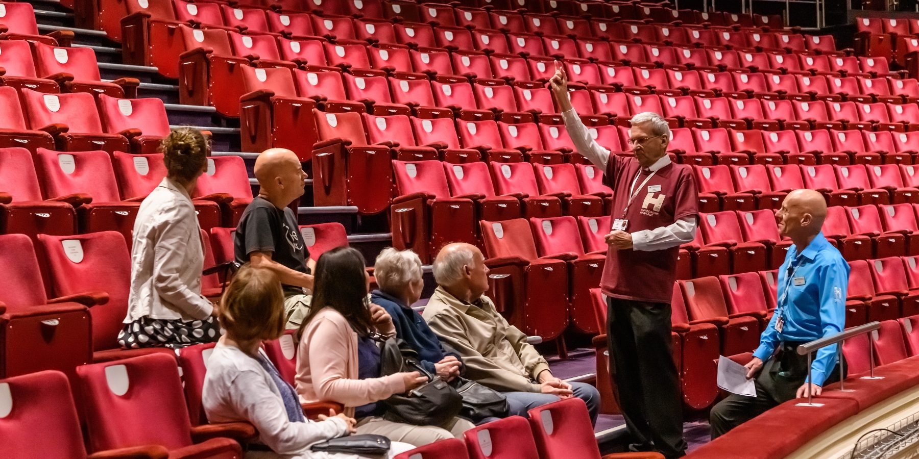 Man points and gesticulates hands whilst standing in auditorium, showing a seated group the surrounding area.