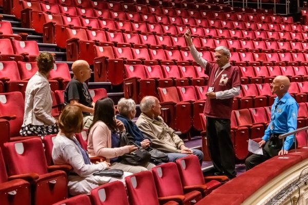 Man points and gesticulates hands whilst standing in auditorium, showing a seated group the surrounding area.