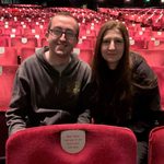 Two people sit together in empty theatre, smiling at the camera.