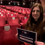 Person sitting in empty theatre smiles while holding certificate with Name a Seat information.