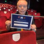 Man sits looking at camera while holding certificate detailing Name a Seat information.