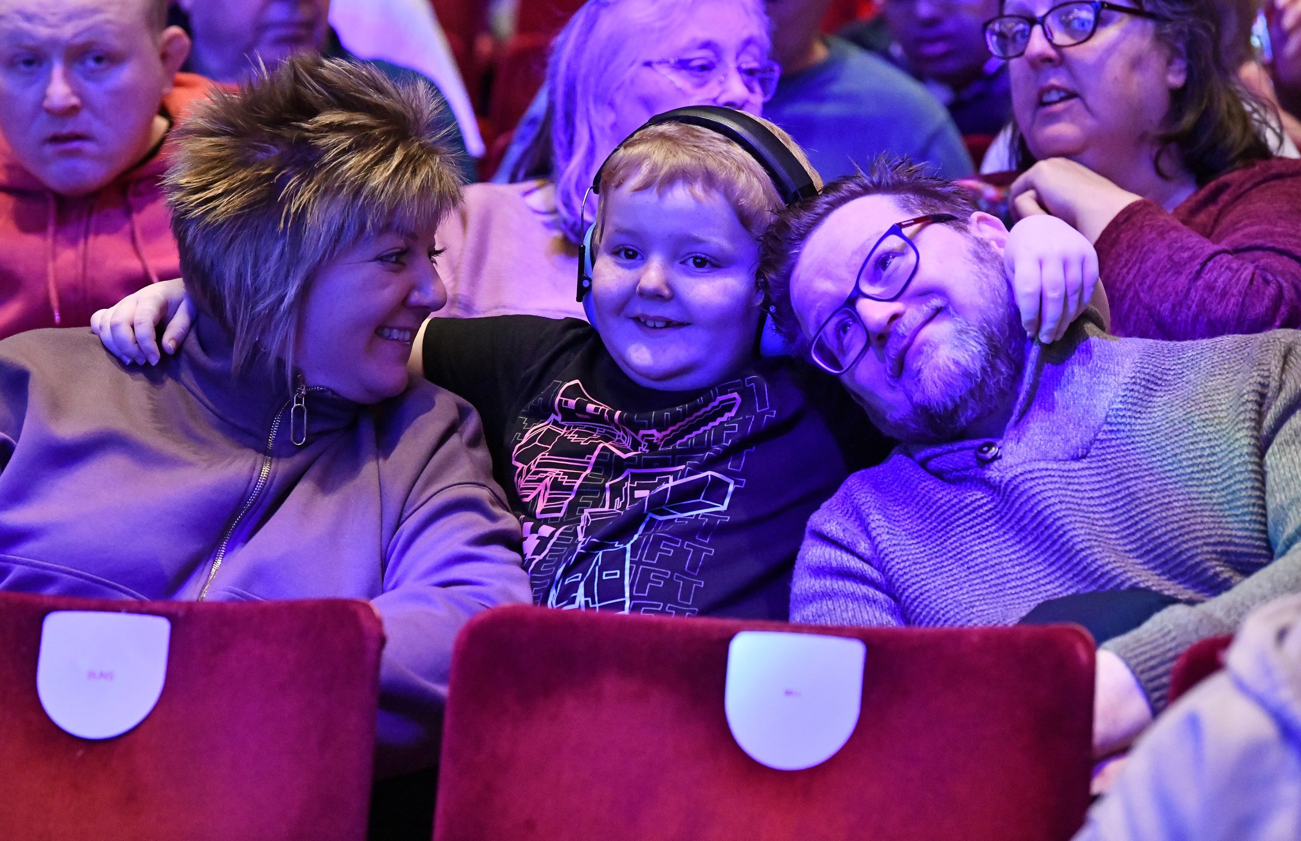 Two adults sit either side of a child wearing noise-reducing headphones while watching a performance.