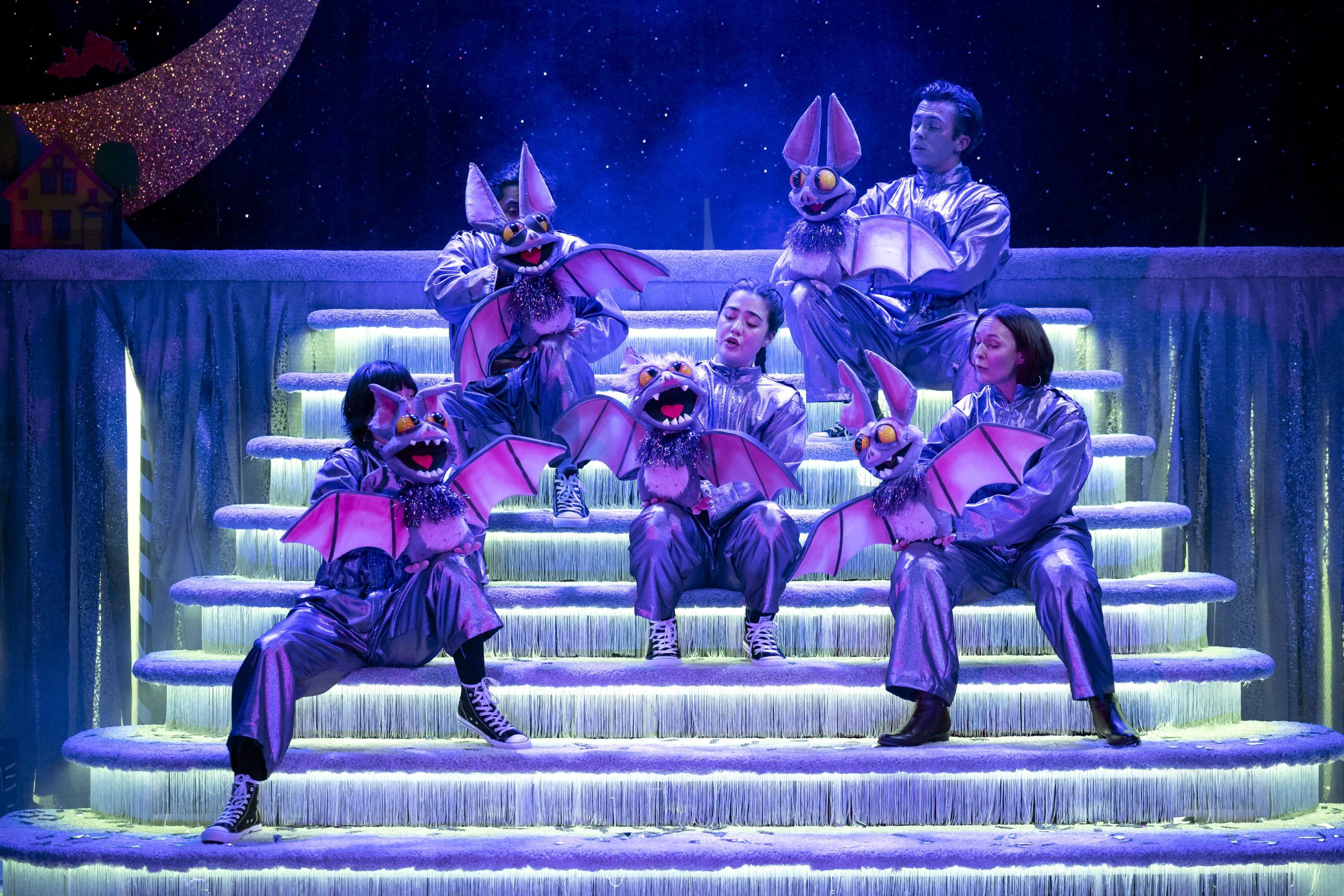 Five performers from The Jingleclaw musical sit on stairs on stage holding puppet bats.