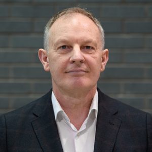 Man in front of grey brick wall wearing a white shirt and dark blazer.