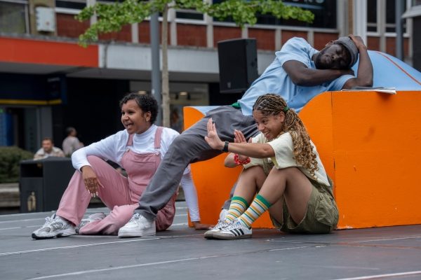 A man (performer) reclines as if asleep on a large, orange and blue prop sofa. His grandchildren (performers) sit at on the floor near him.