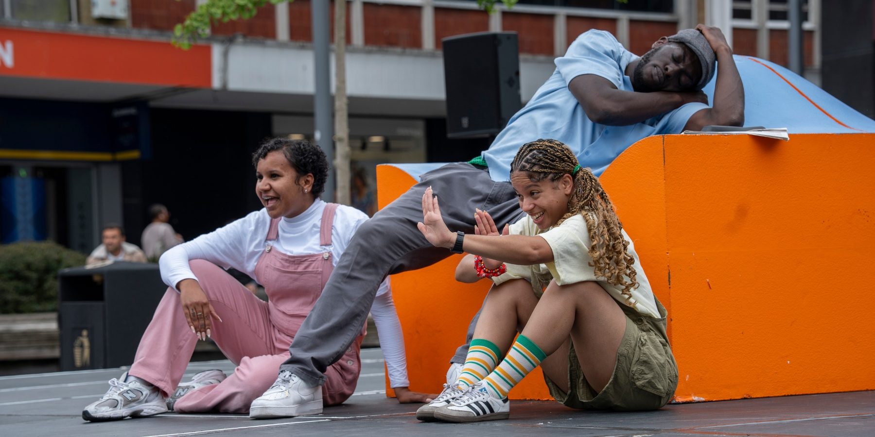 A man (performer) reclines as if asleep on a large, orange and blue prop sofa. His grandchildren (performers) sit at on the floor near him.