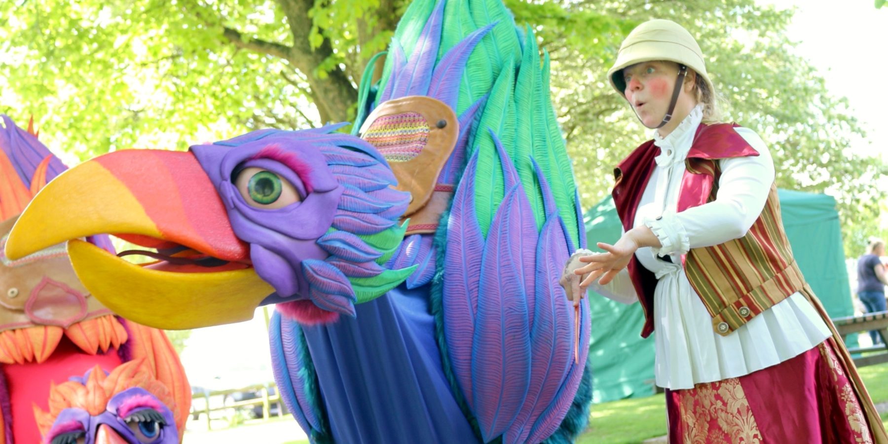 A large, colourful bird-like puppet being moved by a performer wearing an explorer's outfit.