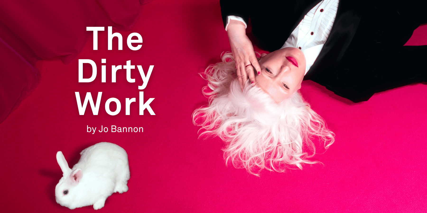 A photograph of Jo Bannon lying on a vibrant pink carpet with her white hair fanned out around her head. She is wearing a black suit and white shirt and hot pink lipstick. Her hand is held to her forehead. A small white rabbit is positioned nearby. The background features a rich pink curtain.