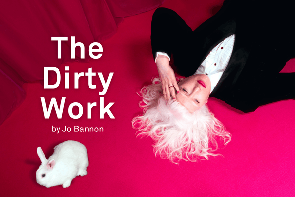 A photograph of Jo Bannon lying on a vibrant pink carpet with her white hair fanned out around her head. She is wearing a black suit and white shirt and hot pink lipstick. Her hand is held to her forehead. A small white rabbit is positioned nearby. The background features a rich pink curtain.